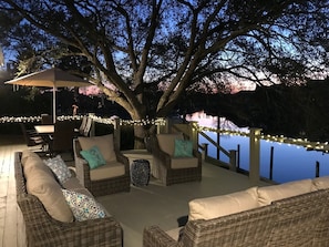 An outdoor patio with wicker furniture, string lights, and a view of a body of water at dusk. - French Quarter Retreat 112 / Spacious Waterfront Home Near New Orleans (Slidell)