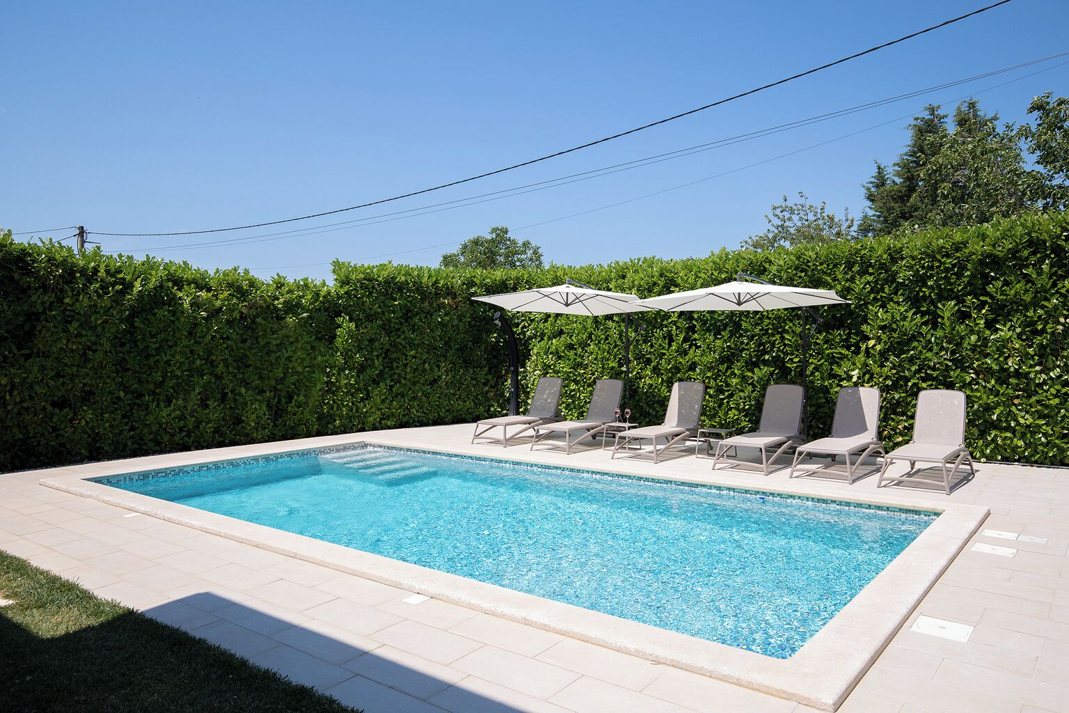 A rectangular swimming pool with a tiled edge, surrounded by a low hedge, and equipped with six lounge chairs and two umbrellas.