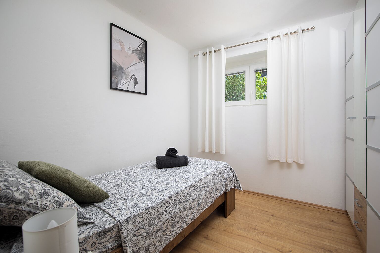 Een slaapkamer met een bed, een raam met gordijnen, een kledingkast en een lijst met een afbeelding aan de muur.