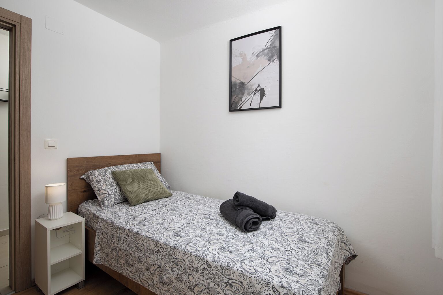 Uma cama de solteiro com um cobreleito estampado, um travesseiro verde e uma toalha enrolada no chão. Tem uma mesa de cabeceira com uma luminária e um quadro pendurado na parede.