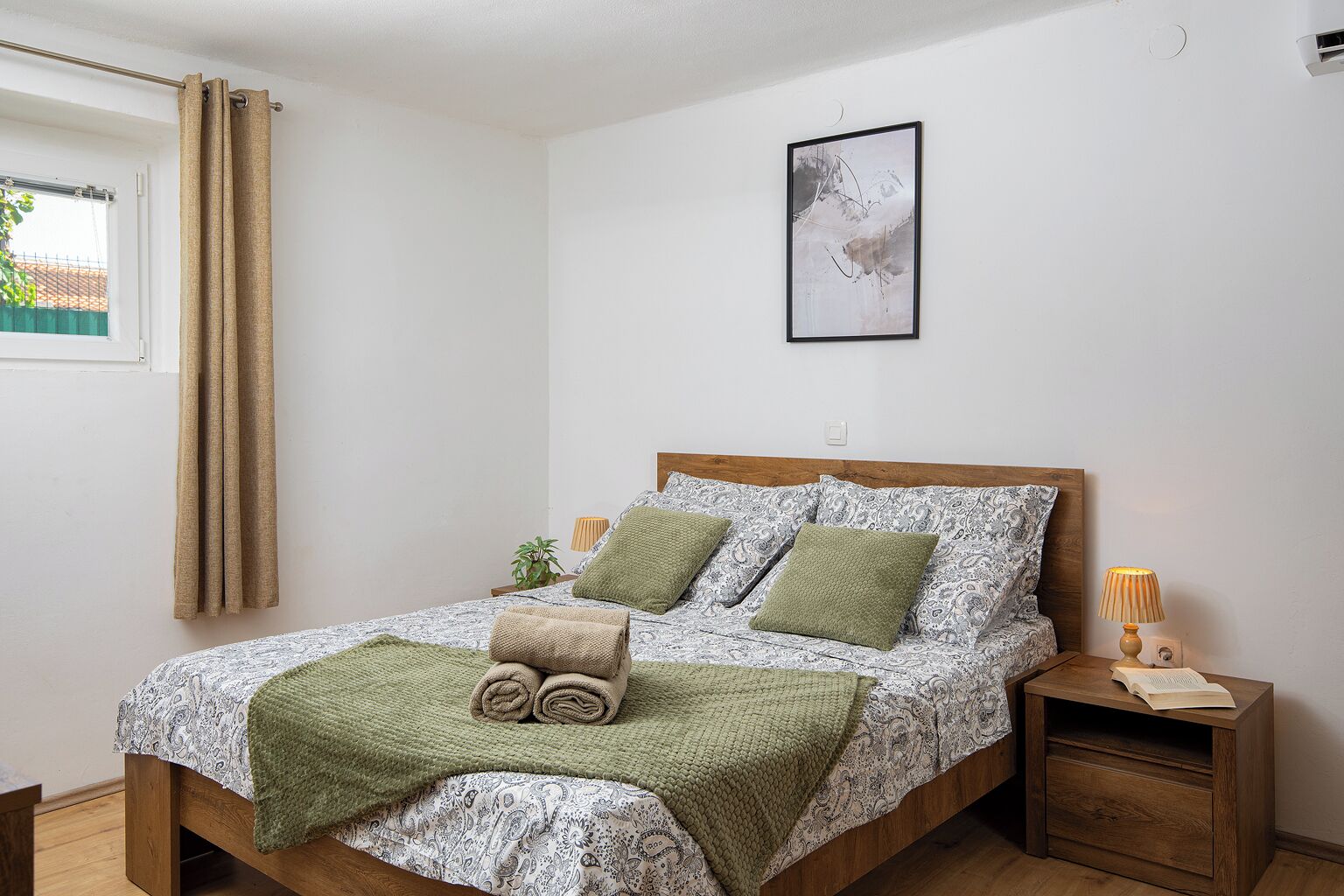 A bedroom with a bed, a bedside table with a lamp, a framed picture on the wall, and a window with curtains.