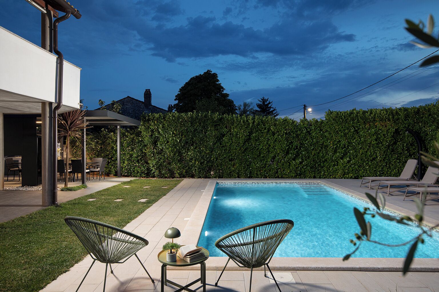 A swimming pool with lounge chairs, a table with a lamp and book, and a hedge in the background.