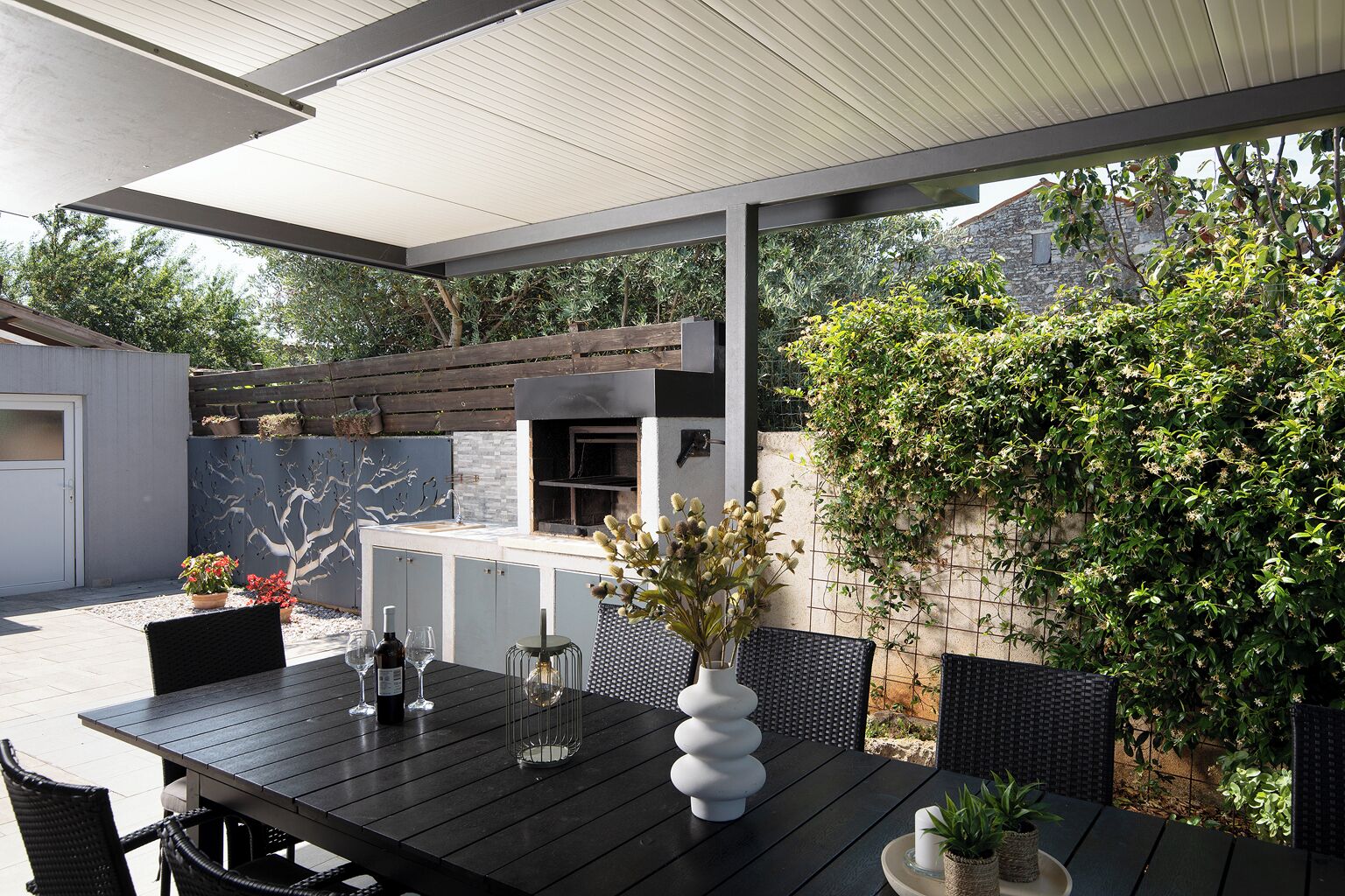 Eine Terrasse im Freien mit Esstisch, integriertem Grill und einer Steinwand mit Kletterpflanzen.
