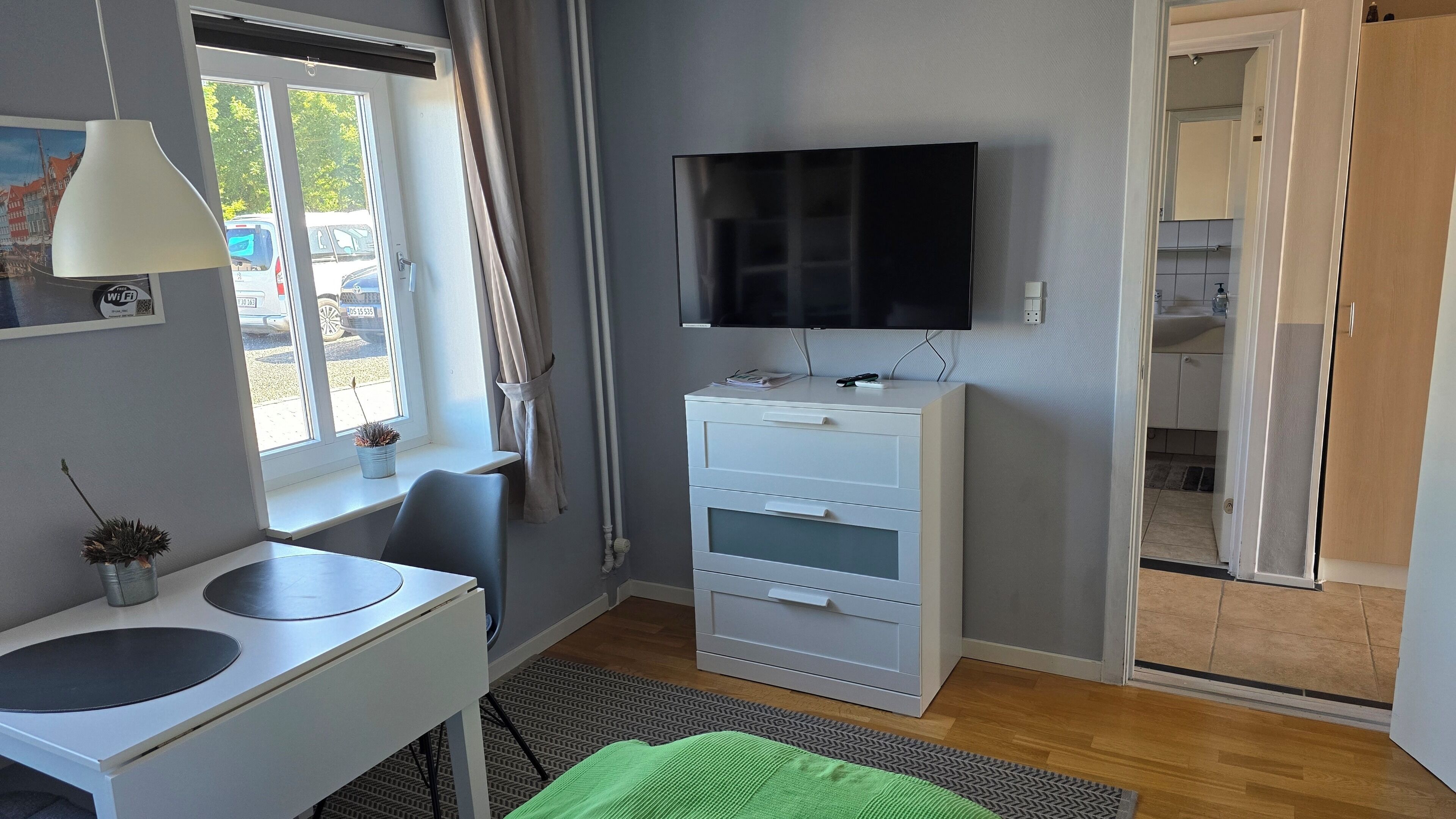 A room with a desk, chair, TV, storage unit, and a window with curtains.