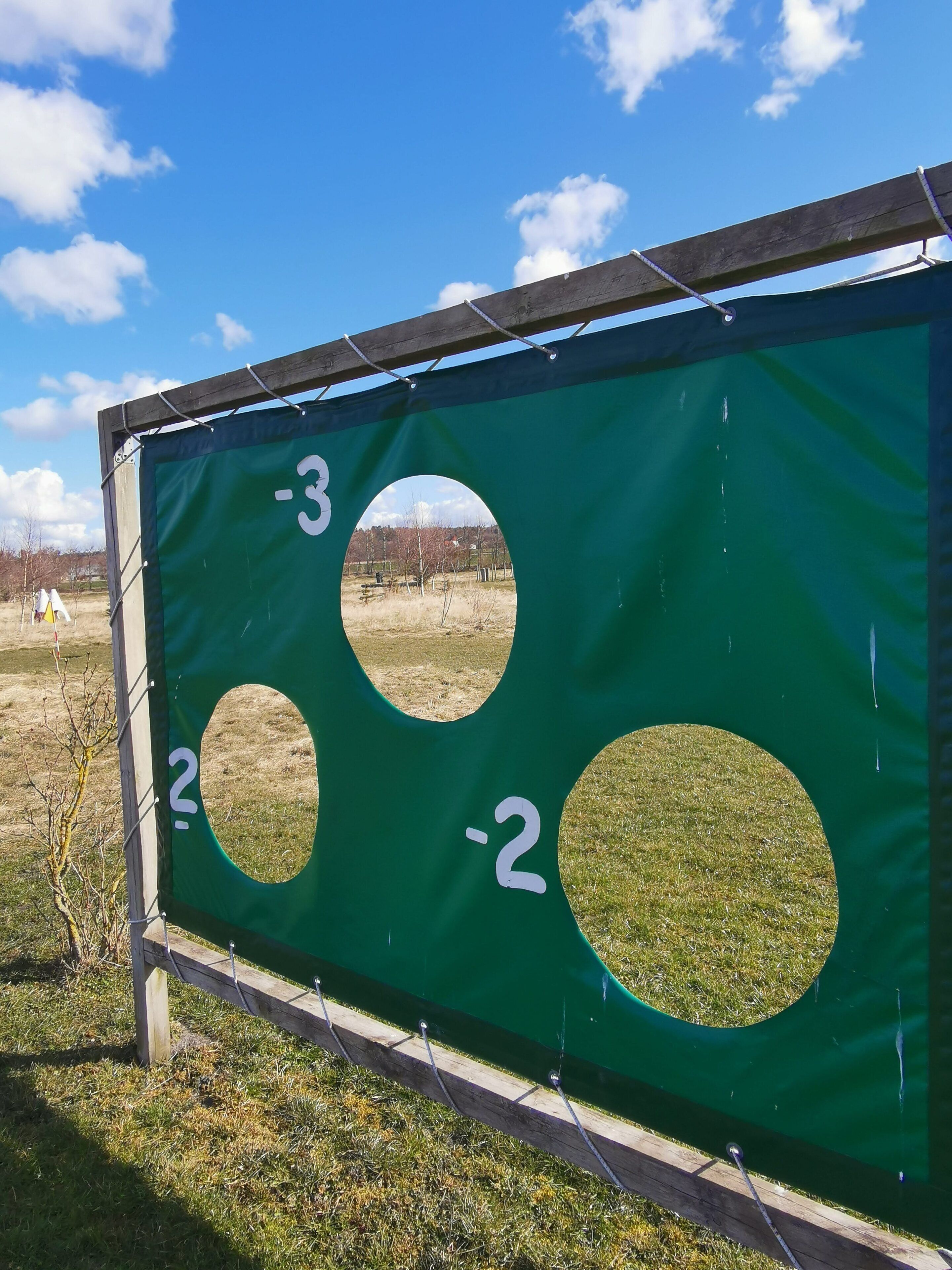 Un tablero de tiro al blanco verde con tres agujeros numerados, sobre un cielo azul despejado.