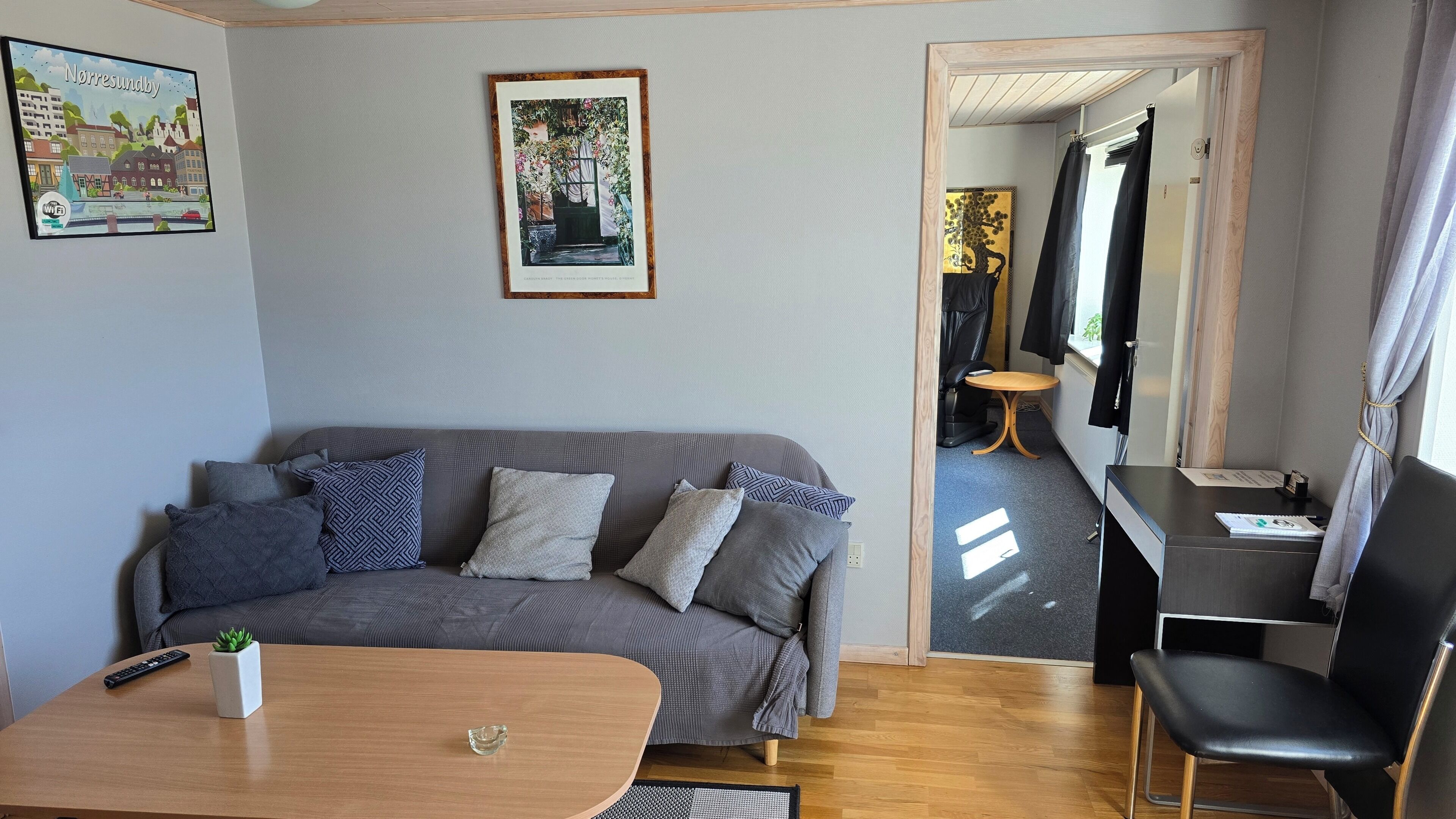 A room with a grey sofa, a small table with a plant, a desk with a chair, and a painting on the wall.