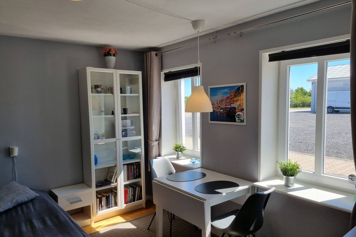Quarto moderno com cama, escrivaninha com cadeira, estante, janela com vista para um prédio e luminária pendente.
