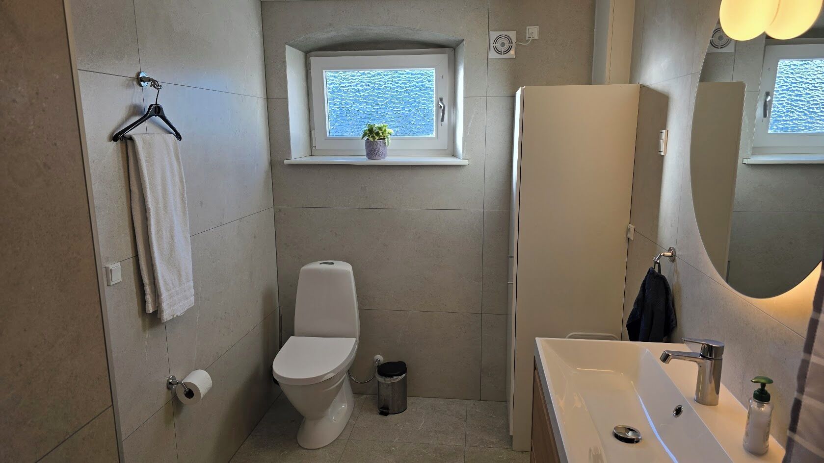 Une salle de bain avec des toilettes, un lavabo, une fenêtre avec une plante et un miroir.