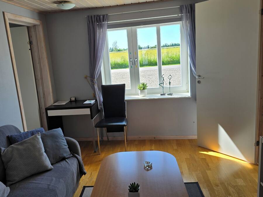 A room with a grey sofa, a black chair, a wooden table, a desk, and a window with a view of a field.