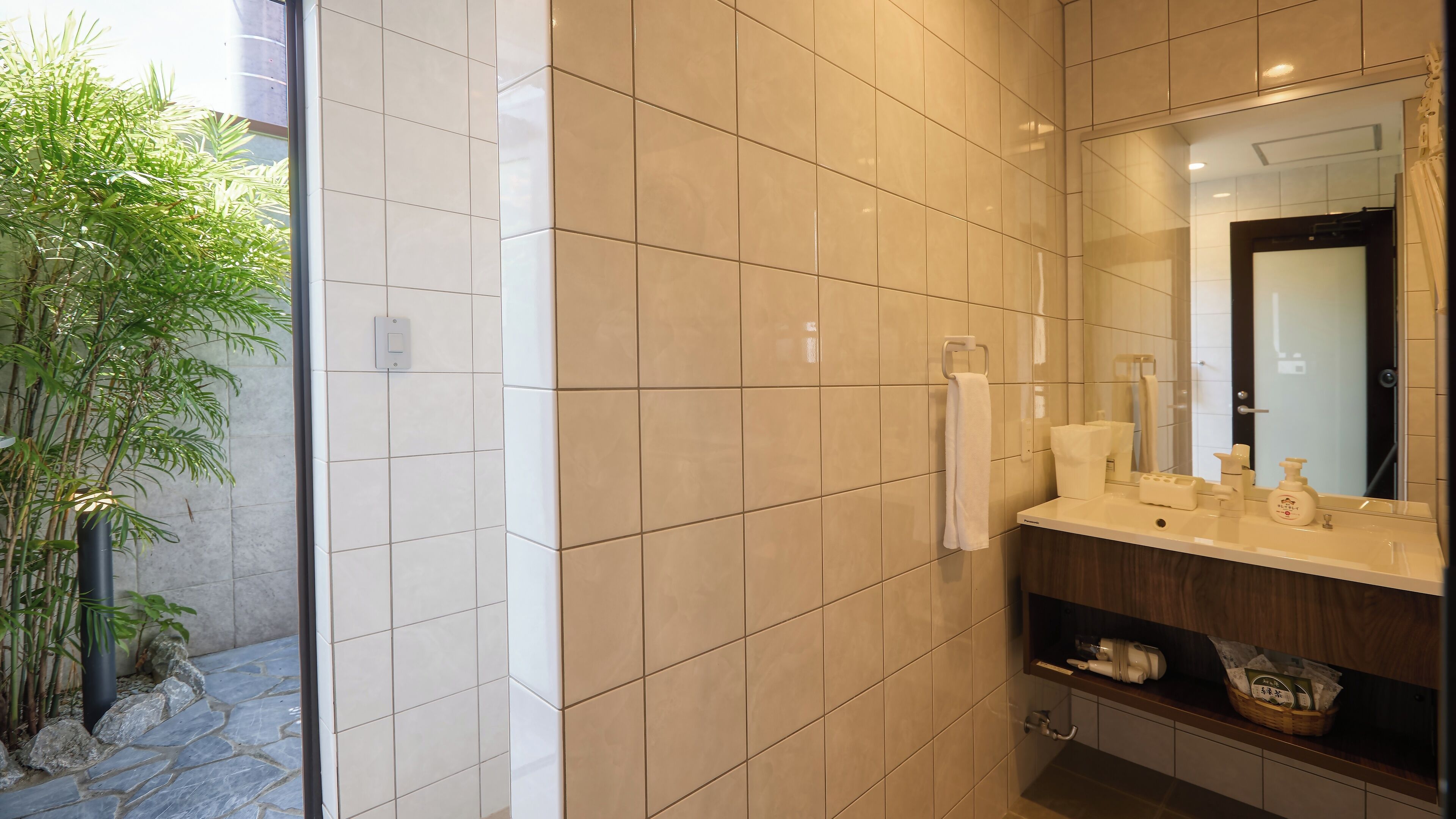 A modern bathroom with beige tiles, a wooden vanity, a mirror, and a bamboo plant.