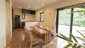 A dining area with a wooden table and chairs, a kitchen in the background, and a large window with a view of greenery. - RERESO Okinawa churaumi (Motobu)