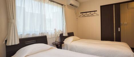 A hotel room with two beds, a window with sheer curtains, an air conditioning unit, a bedside table with a vase of flowers, and a coat rack with hangers.