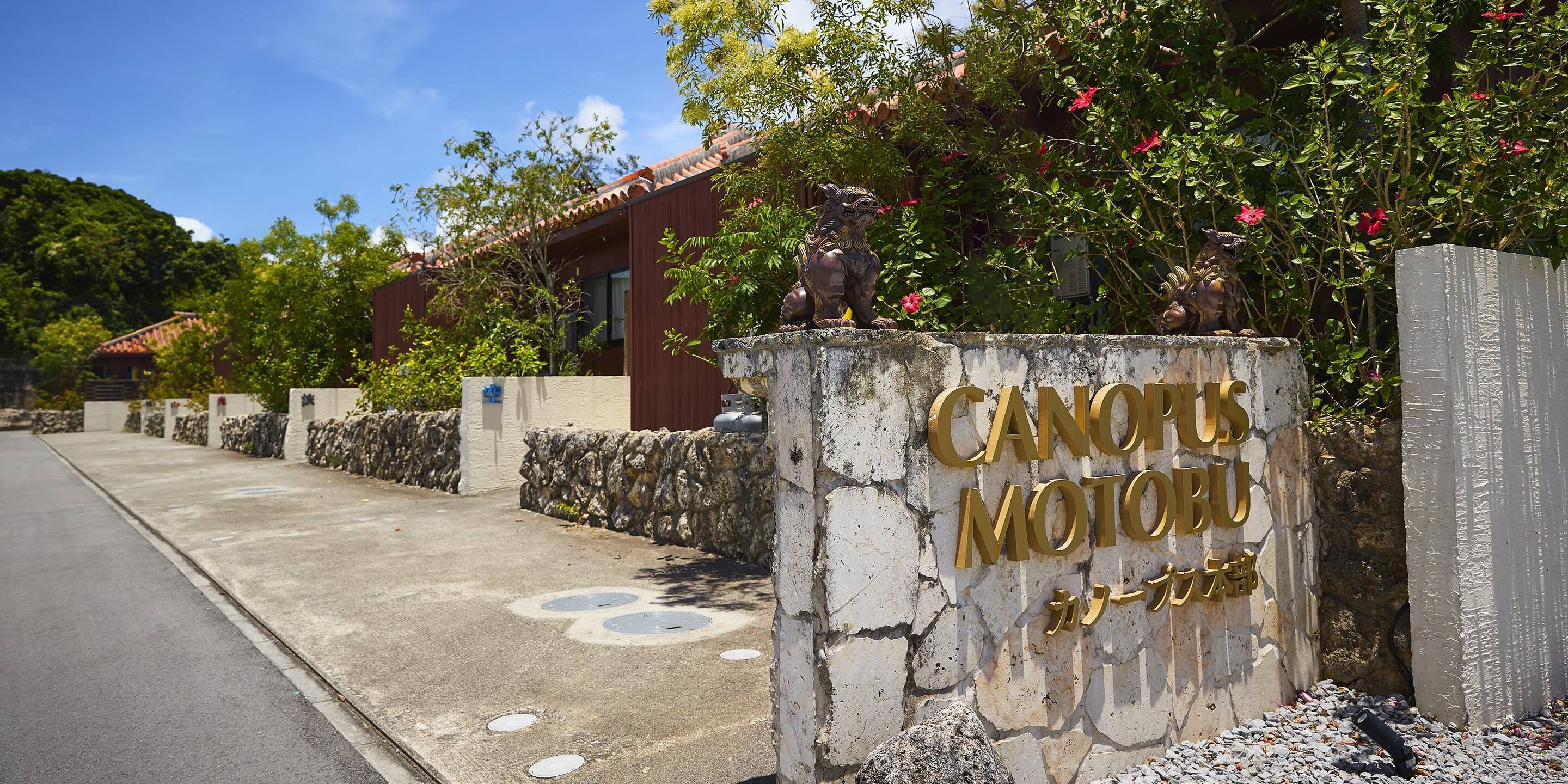 A building entrance with a stone wall and a metal sign reading "CANOPOS MOTOBU".