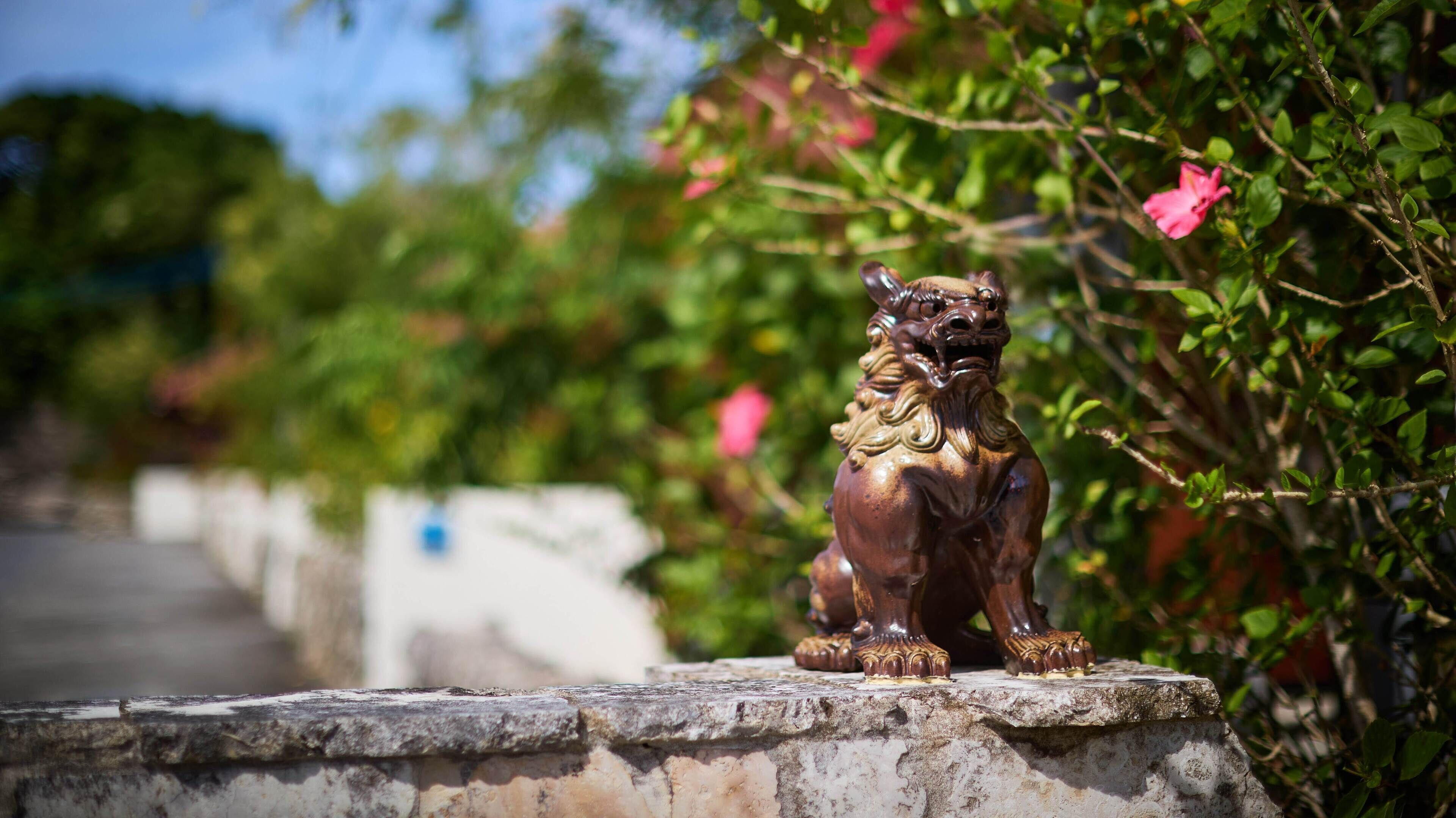 Una estatua de bronce de un león sobre un pedestal de piedra rodeado de follaje y flores rosas.