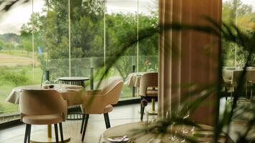 Ein Essbereich mit Tischen für zwei Personen, große Fenster mit Blick ins Freie und eine Pflanze im Vordergrund.
