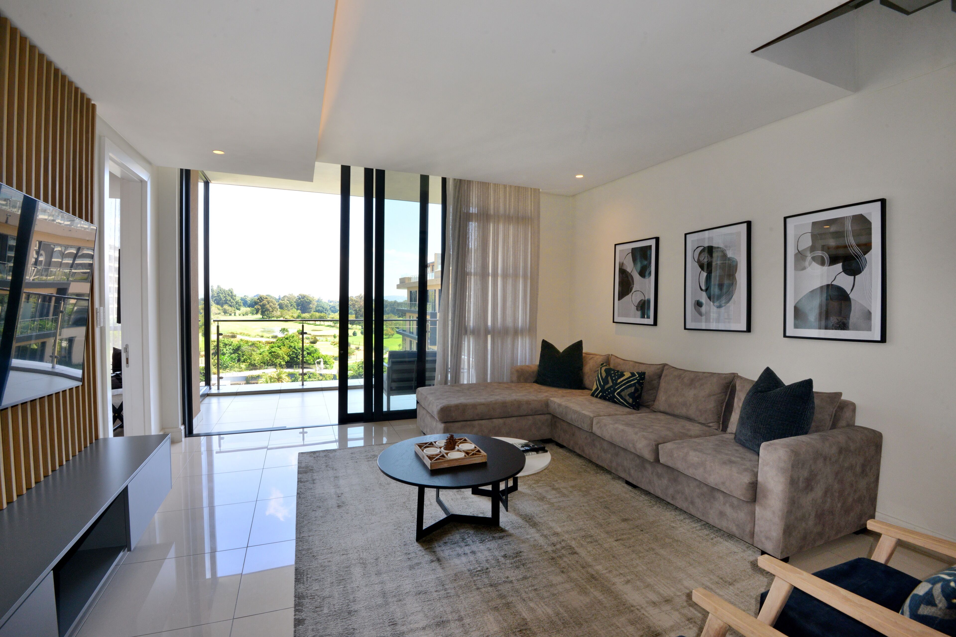 Un salon moderne avec un grand canapé d’angle, une table basse et un téléviseur à écran plat. De grandes fenêtres offrent une vue sur la verdure environnante.