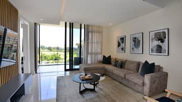 Un salon moderne avec un grand canapé d’angle, une table basse et un téléviseur à écran plat. De grandes fenêtres offrent une vue sur la verdure environnante.