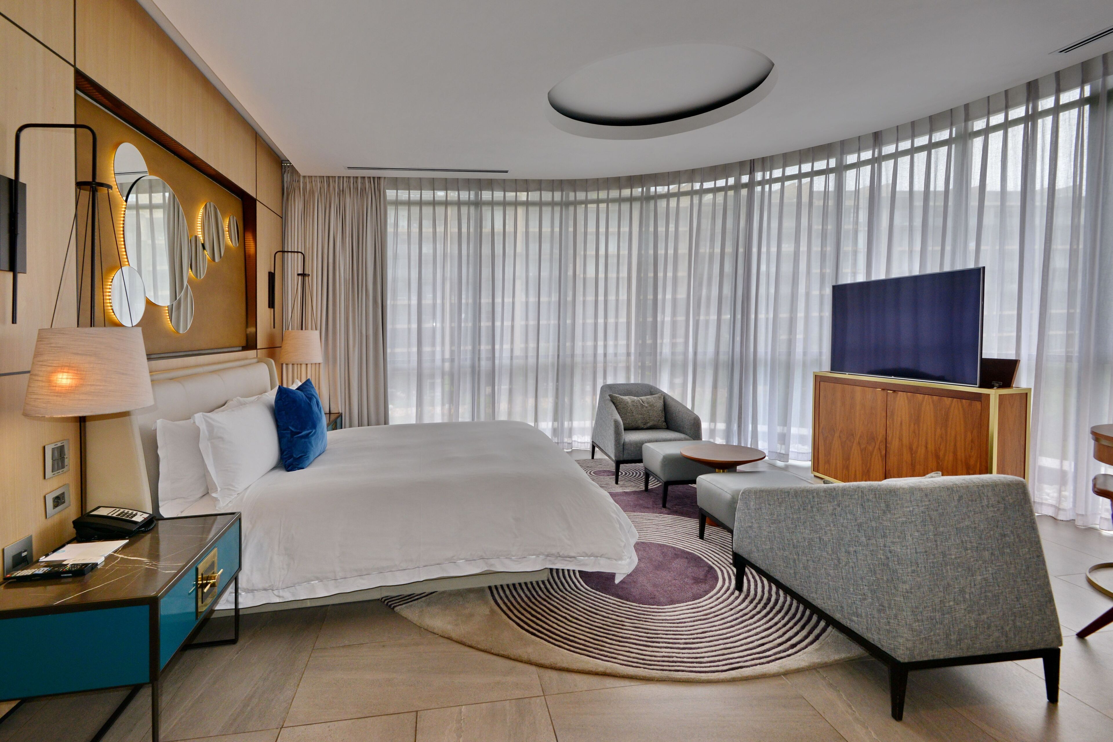 Habitación de hotel moderna con una cama grande, un escritorio con lámpara, televisión y vistas a la ciudad a través de amplios ventanales.