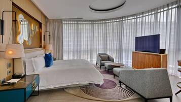 Habitación de hotel moderna con una cama grande, un escritorio con lámpara, televisión y vistas a la ciudad a través de amplios ventanales.
