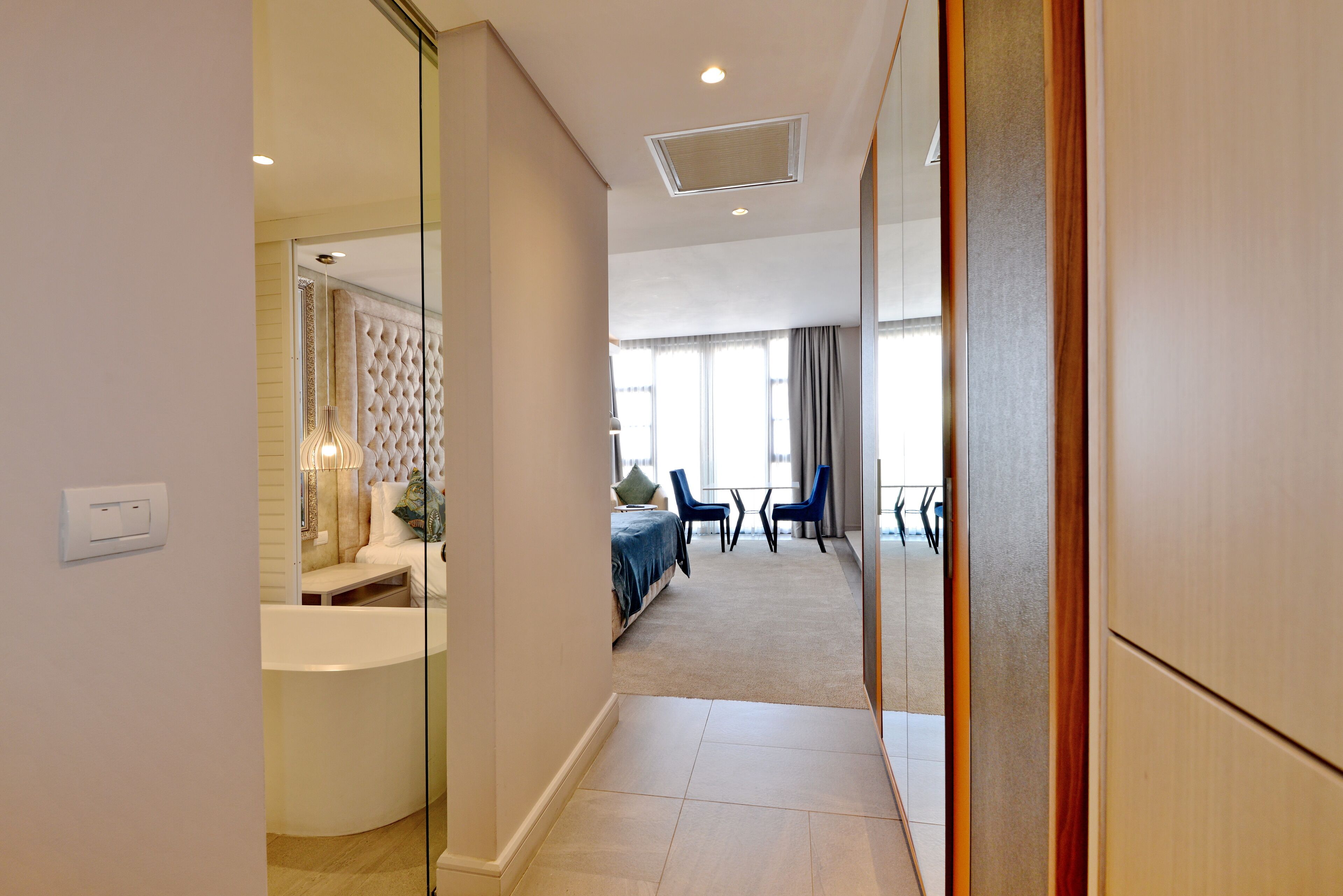 A hotel room with a curved bathtub, a tufted headboard, a seating area with a sofa and chairs, and a large window with curtains.