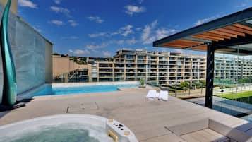 A rooftop pool area with a hot tub, lounge chairs, and a view of a hotel building.