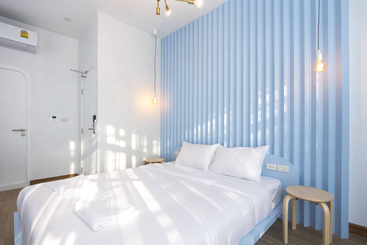 A modern bedroom with a large bed, a bedside table, a hanging light fixture, and a window with vertical blinds.