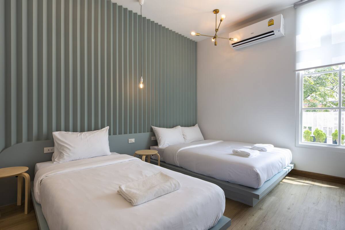A modern hotel room with two beds, a wooden floor, a window with a view, and an air conditioning unit.