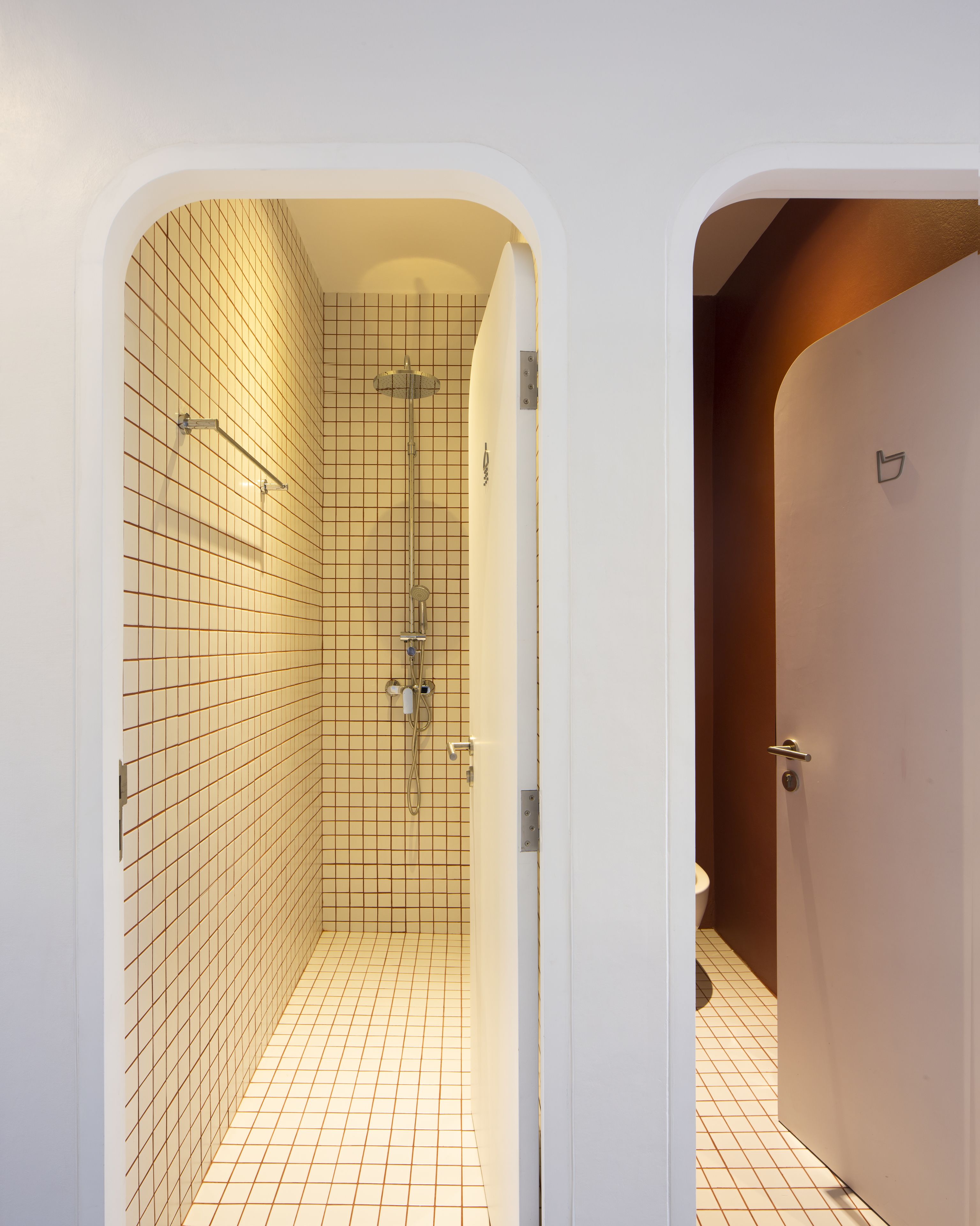 Une petite salle de bain carrelée, équipée d’une douche, de toilettes et d’une porte donnant sur une autre pièce.