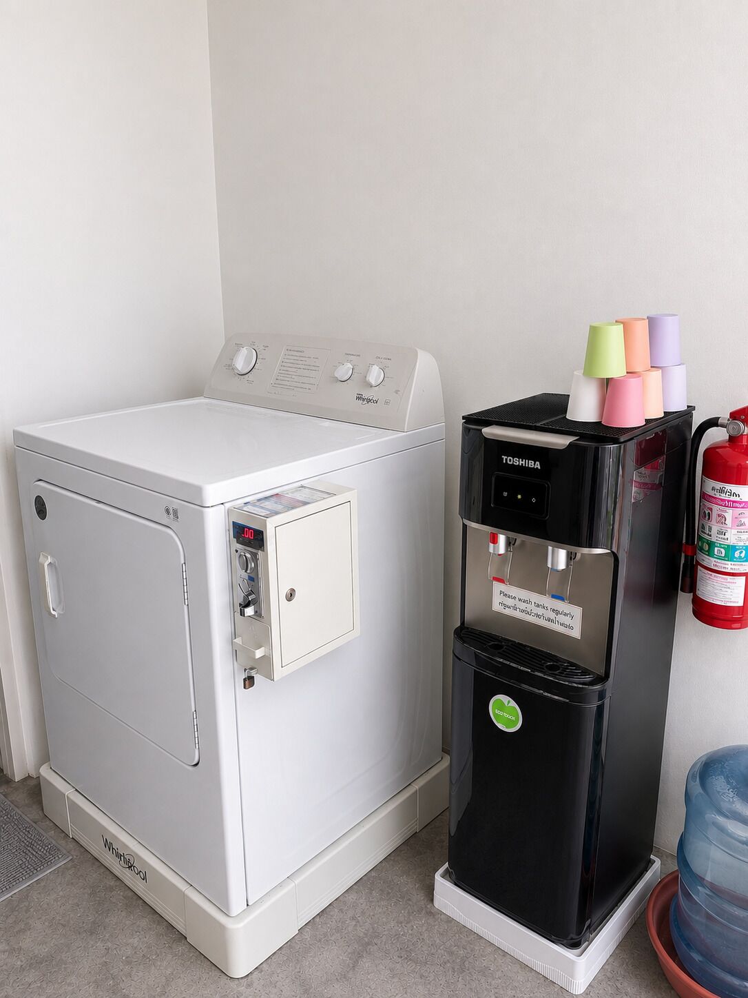 Una lavadora y secadora Whirlpool, una cafetera Toshiba y un dispensador de agua, con un extintor cerca.