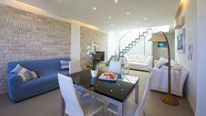 A modern living room with a stone wall, a blue sofa, a white sofa, a glass dining table with chairs, a floor lamp, and a staircase leading to an upper level. - Villa Mondello Addaura Sea View (Palermo)