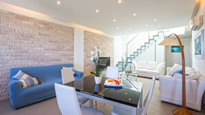 A modern living room with a blue sofa, white armchairs, a glass dining table, and a staircase leading to an upper floor. - Villa Mondello Addaura Sea View (Palermo)