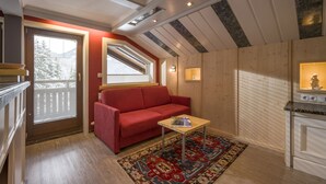 Ein gemütliches Zimmer mit einem roten Sofa, einem kleinen Tisch und einem gemusterten Teppich. Es gibt ein Fenster mit Jalousien, eine Decke mit Holzbalken und eine kleine Küchenzeile mit einer Arbeitsplatte.