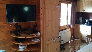 Ein Holzblockhaus-Innenraum mit an einer Wandregal montiertem Fernseher, einer Küchenzeile mit Holzschränken und einem Esstisch mit Stühlen.
