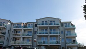 A multi-story residential building with balconies, parking in front, and a clear sky.