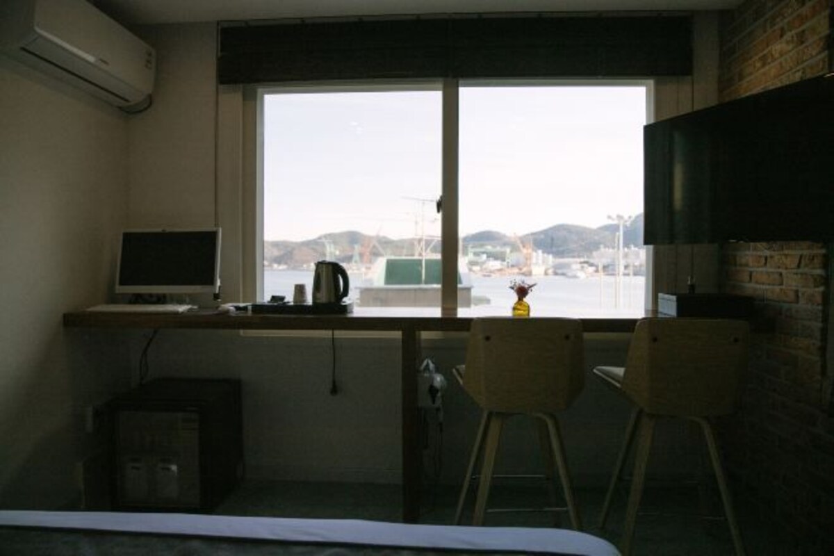 A small room with a desk, a chair, a bed, and a large window with a view of a cityscape.