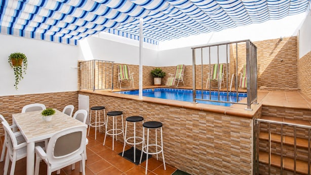 Ein Poolbereich mit Fliesenboden, einem Pool, weißen Barhocker, einem Tisch mit Stühlen und einem gestreiften Sonnensegel.