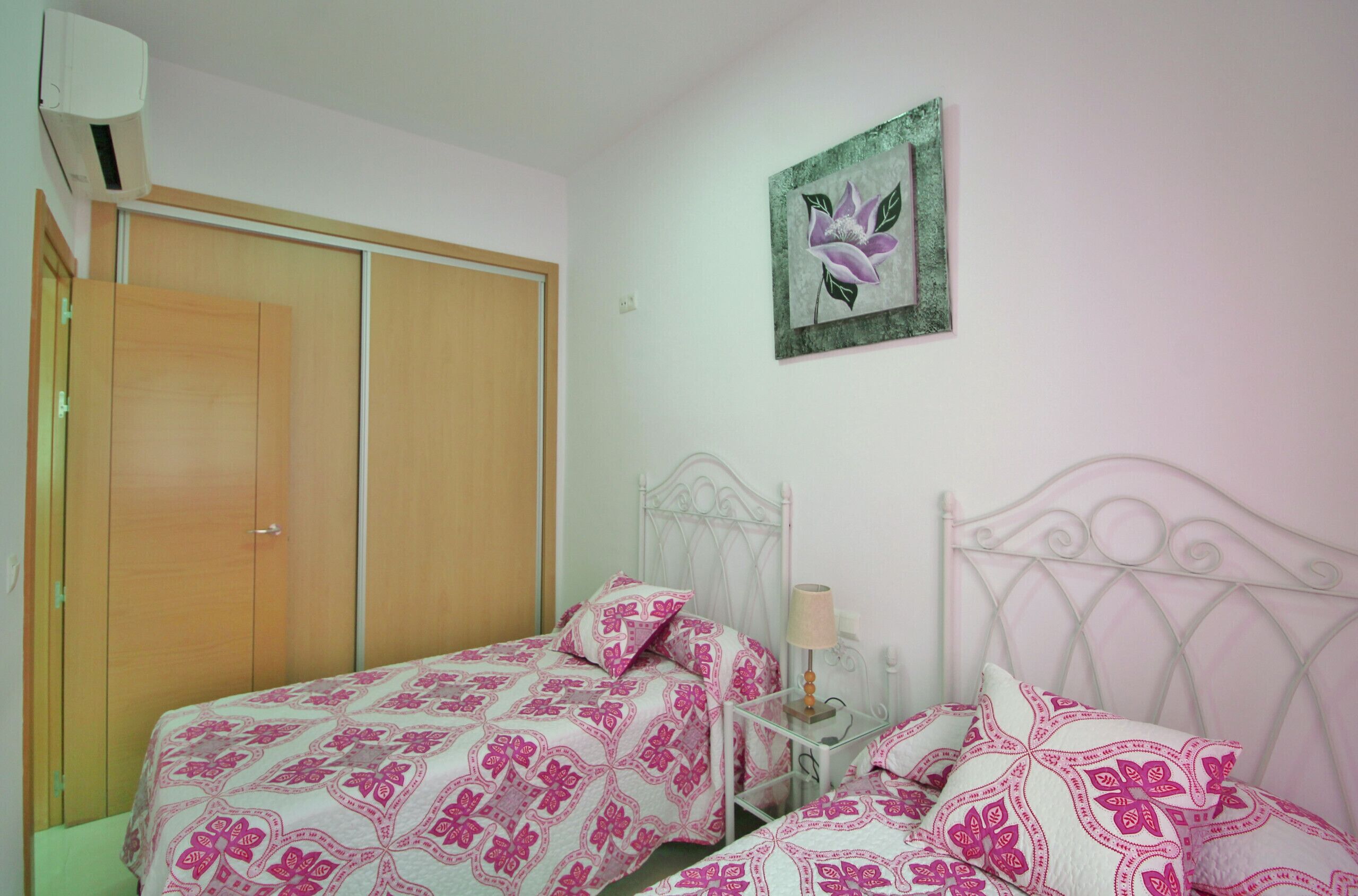 A bedroom with two beds, a wardrobe, a bedside table with a lamp, and a wall-mounted painting.