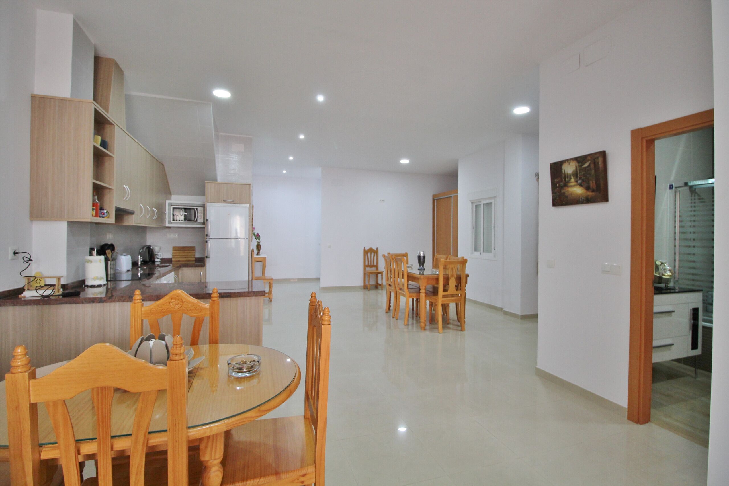 A modern kitchen with white cabinets, a microwave, and a refrigerator. There is a dining area with a round table and wooden chairs. The room has a tiled floor and white walls.