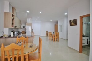 A modern kitchen with wooden cabinets, a microwave, and a refrigerator. There is a dining area with a round glass table and wooden chairs.