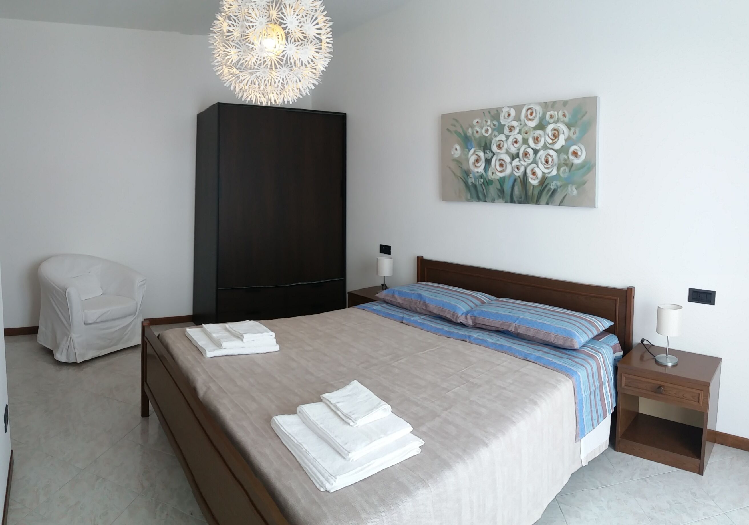 Un lit bien fait, avec des serviettes pliées, une armoire sombre, un tableau de fleurs, une chaise blanche et une table de chevet avec une lampe.