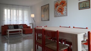A modern living room with a red sofa, a white coffee table, and a dining area with a wooden table and red chairs. There are two paintings on the wall and a floor lamp.