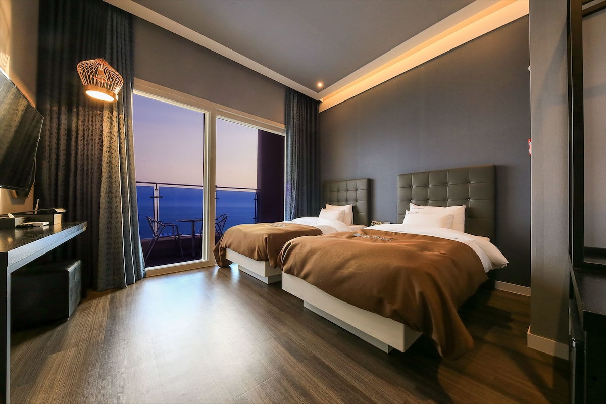 A modern hotel room with two beds, a desk, and a balcony with a view.