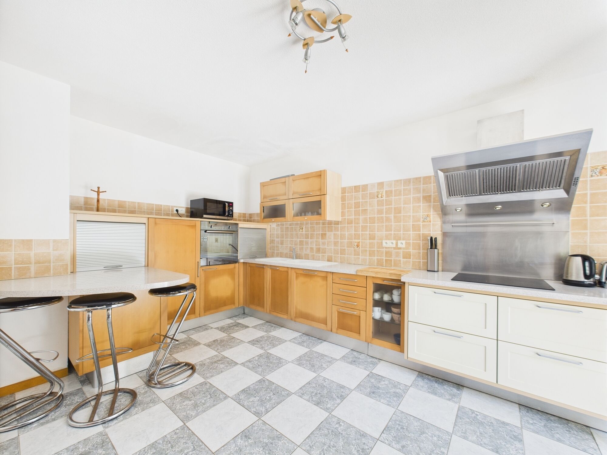 Una cocina moderna con microondas, horno y campana extractora de acero inoxidable. Cuenta con armarios de madera, salpicadero de azulejos y suelo a cuadros.