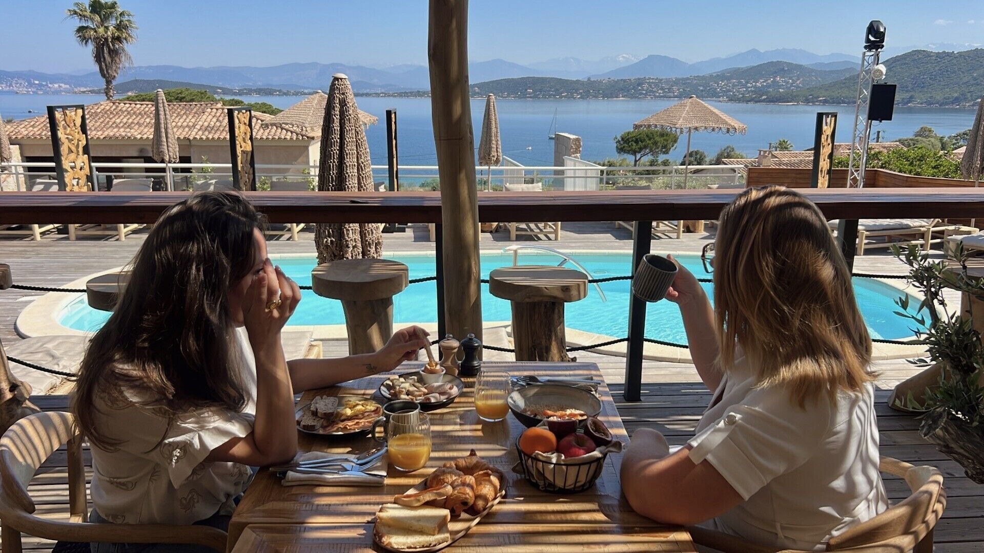 Deux personnes sont assises à une table, dînant, avec une piscine et des montagnes en arrière-plan.