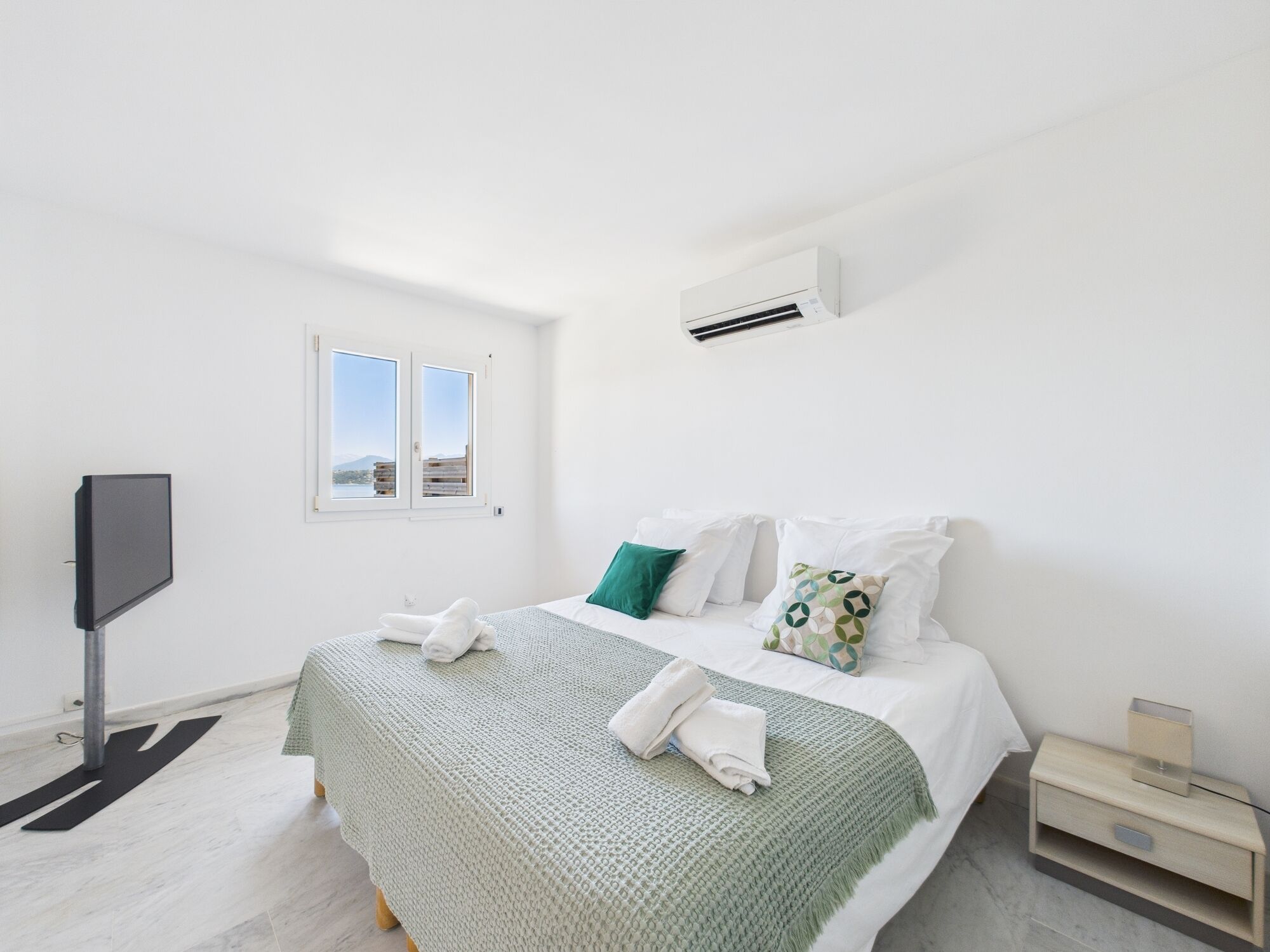Une chambre à coucher avec un lit, des oreillers, une télévision, une fenêtre et un appareil de climatisation.