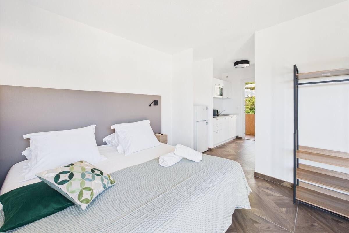 A modern hotel room with a bed, a small kitchenette, and a view of the outdoors.