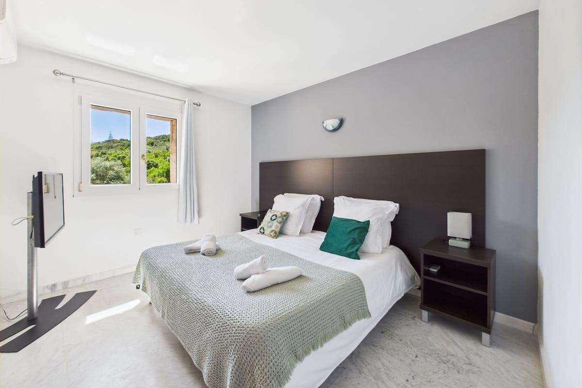 A modern bedroom with a large bed, a bedside table, a nightstand, a TV, and a window with a view of greenery.