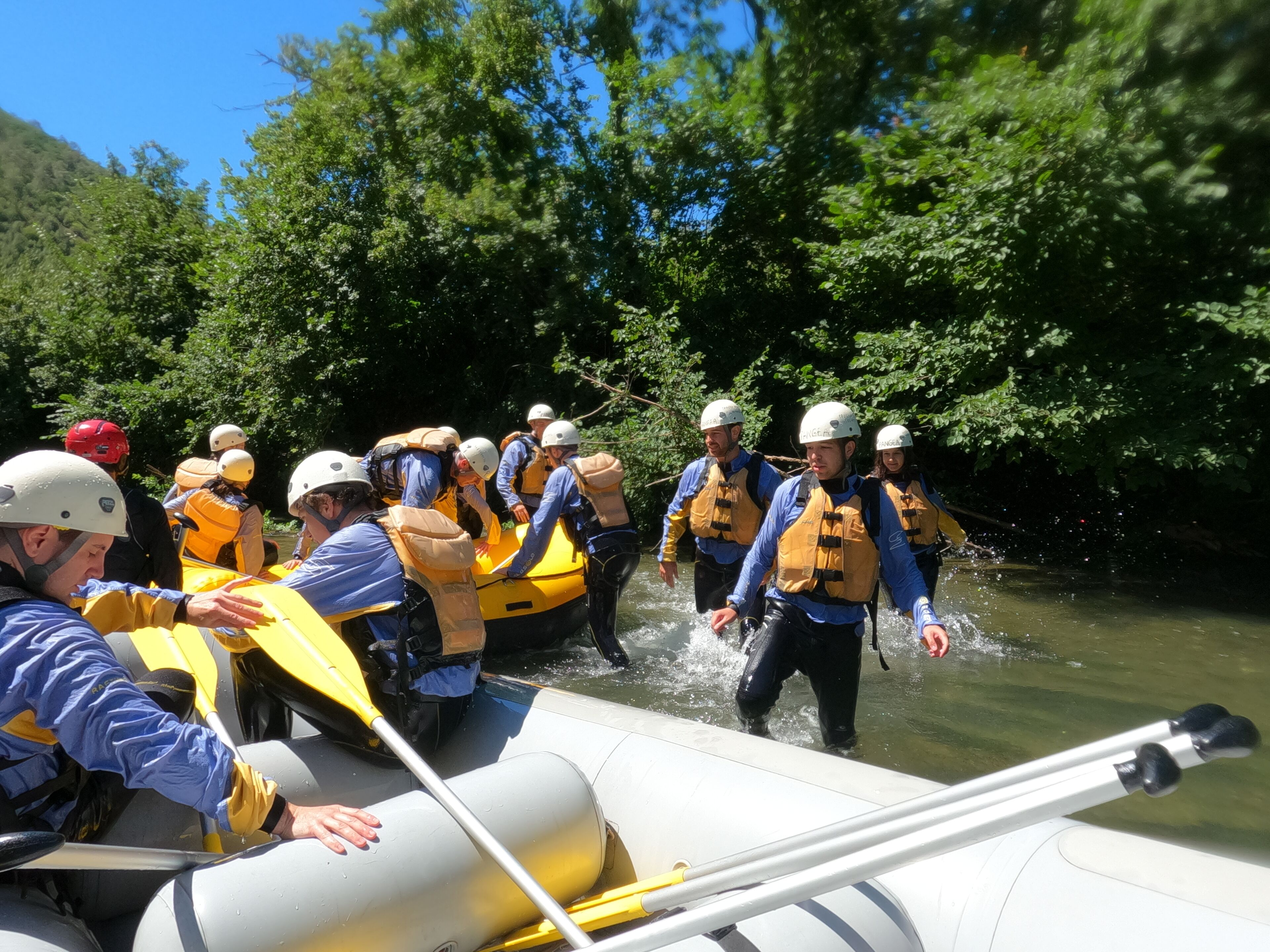 Un groupe de personnes, vêtues de gilets de sauvetage et de casques jaunes, se trouve sur un radeau blanc dans une rivière.