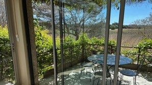 Un porche acristalado con una mesa redonda y sillas, rodeado de vegetación y vistas a un viñedo.