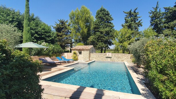 Piscina rectangular con muro de piedra y un pequeño edificio de piedra, rodeada de exuberante follaje y árboles.